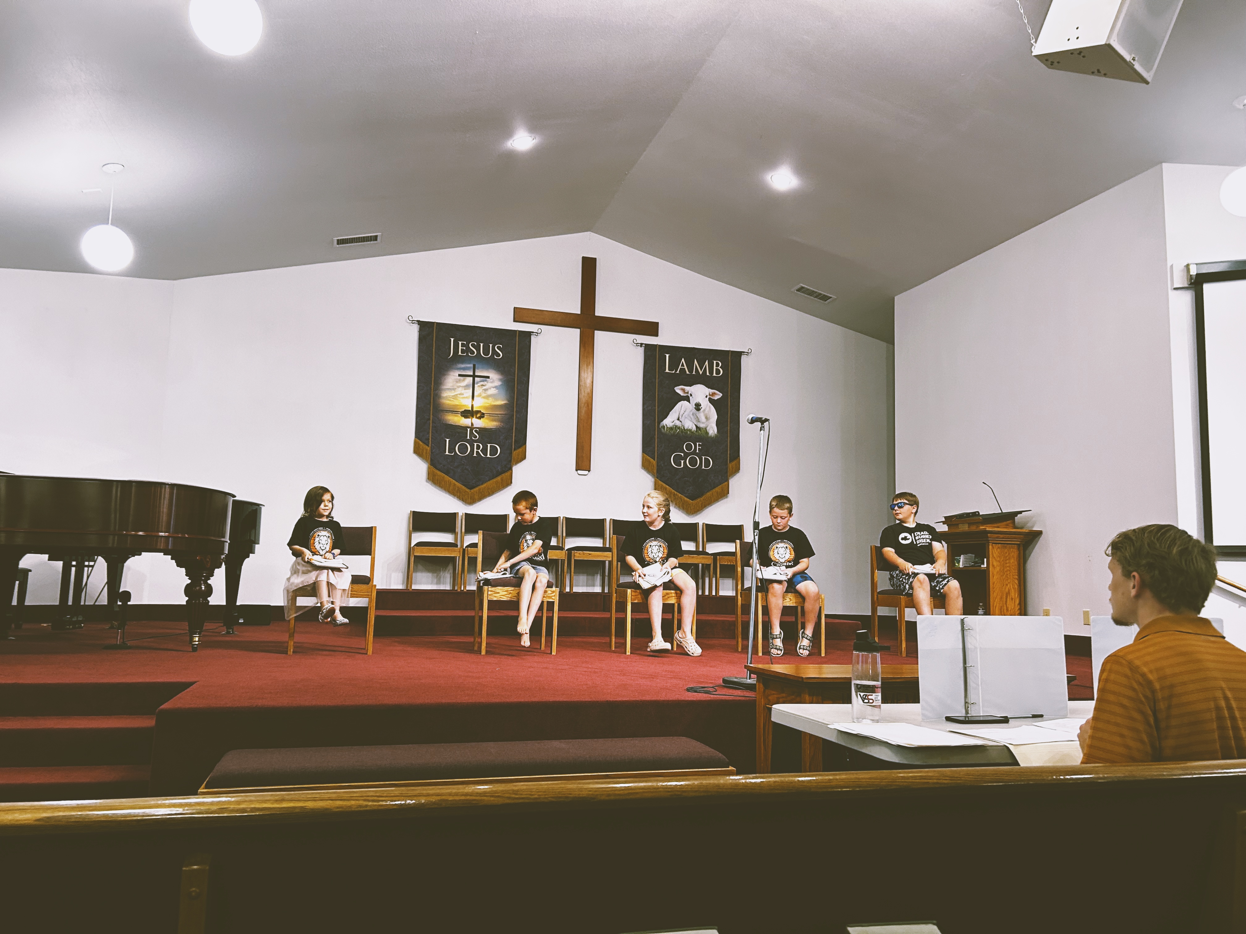 Five children sit on the platform of Valley Baptist Church during a service, with banners reading Jesus is Lord and Lamb of God flanking a wooden cross behind them