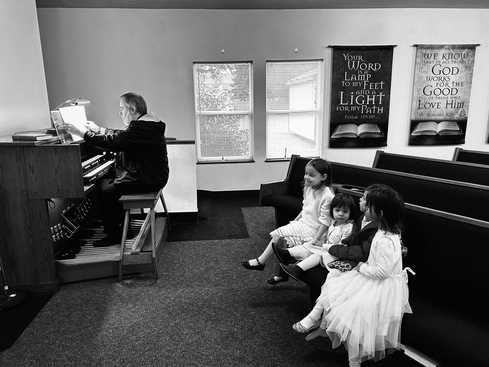 An organist plays at Valley Baptist Church while three young girls watch quietly from the pew, scripture banners on the wall behind